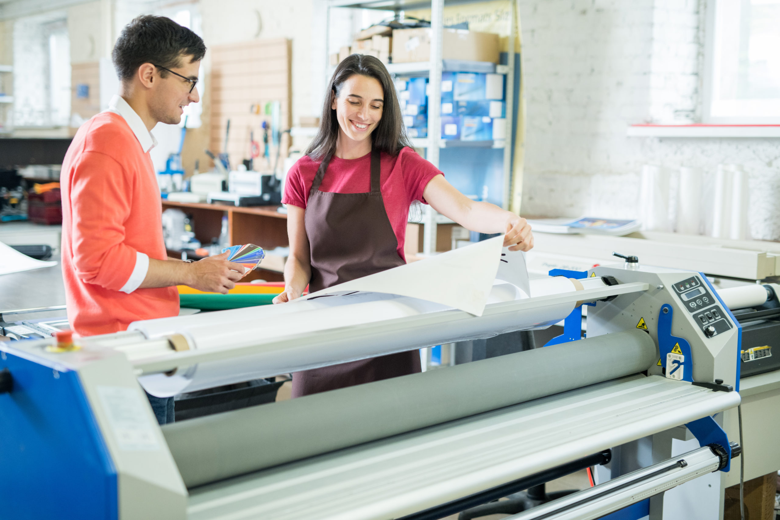 Cheerful excited beautiful lady in apron showing printed banner to manager who picking out color on swatch for printing